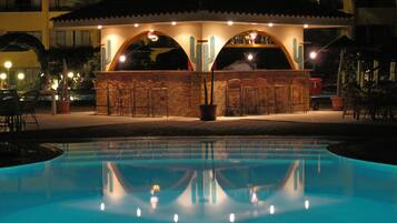 Un espace bar situé au bord de la piscine, avec la piscine en premier plan, entouré de chaises longues et d’un espace de détente couvert doté d’un toit en tuiles.