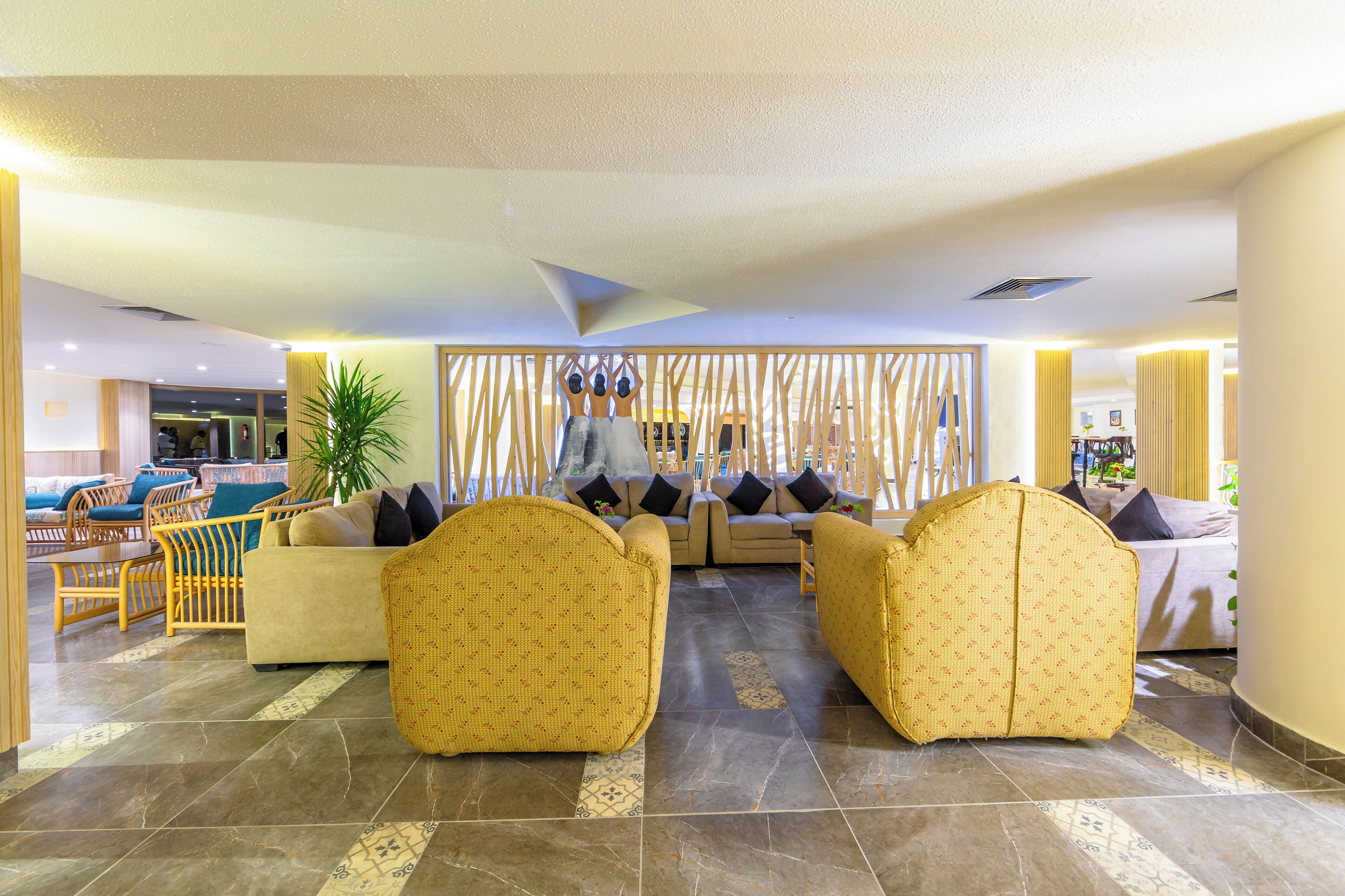 Un hall d’entrée spacieux au design moderne, comprenant un espace de détente central avec des canapés et des fauteuils, une cloison décorative en bois et une grande plante en pot.