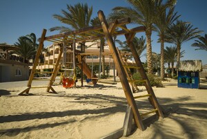 A wooden swing set and a ladder on a sandy beach with palm trees and a building in the background.