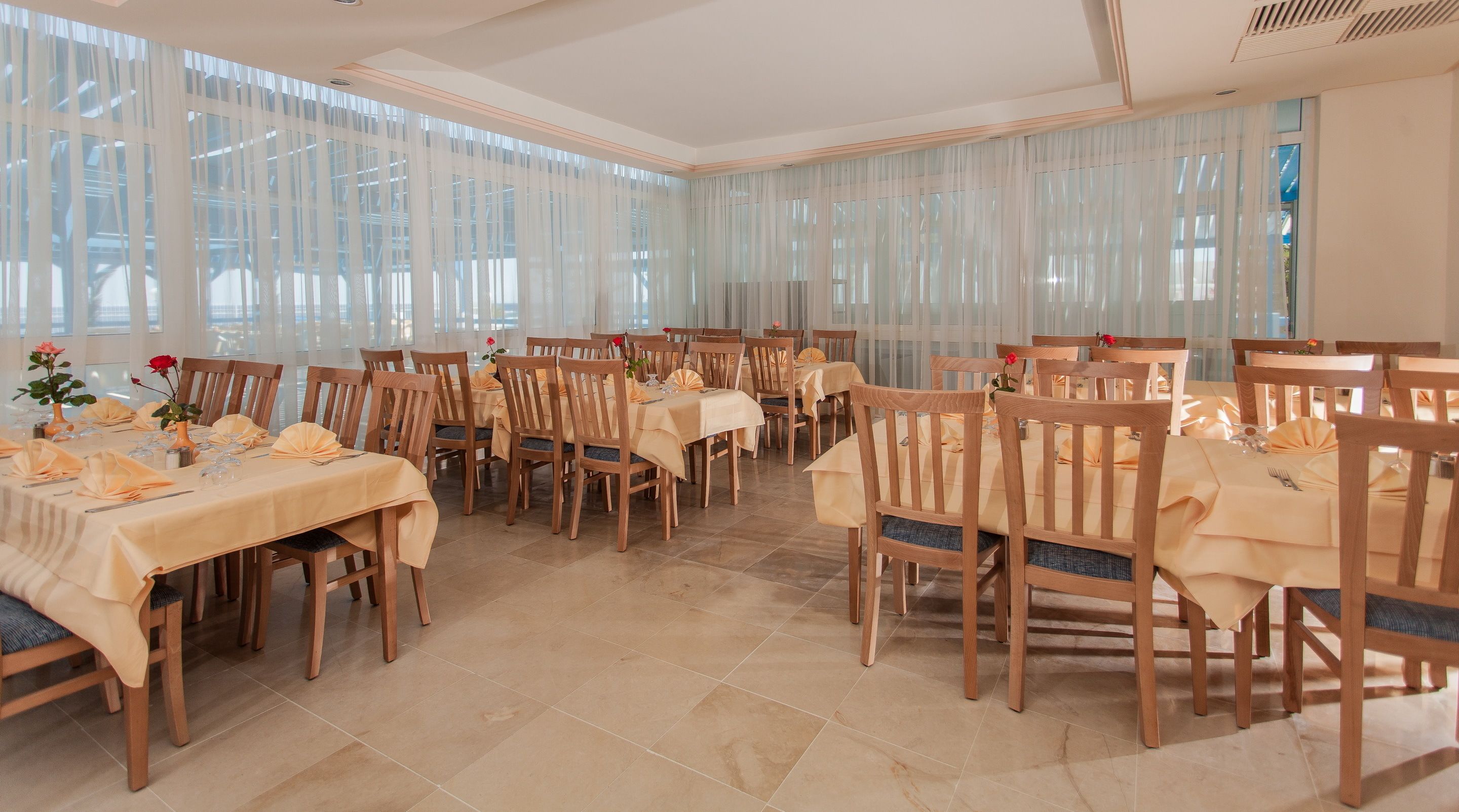 A spacious dining area with multiple tables set for a meal, featuring wooden chairs and a tiled floor.