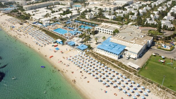 Vue aérienne d’un complexe hôtelier de bord de mer, avec plusieurs piscines, des parasols de plage et un grand bâtiment d’hôtel.