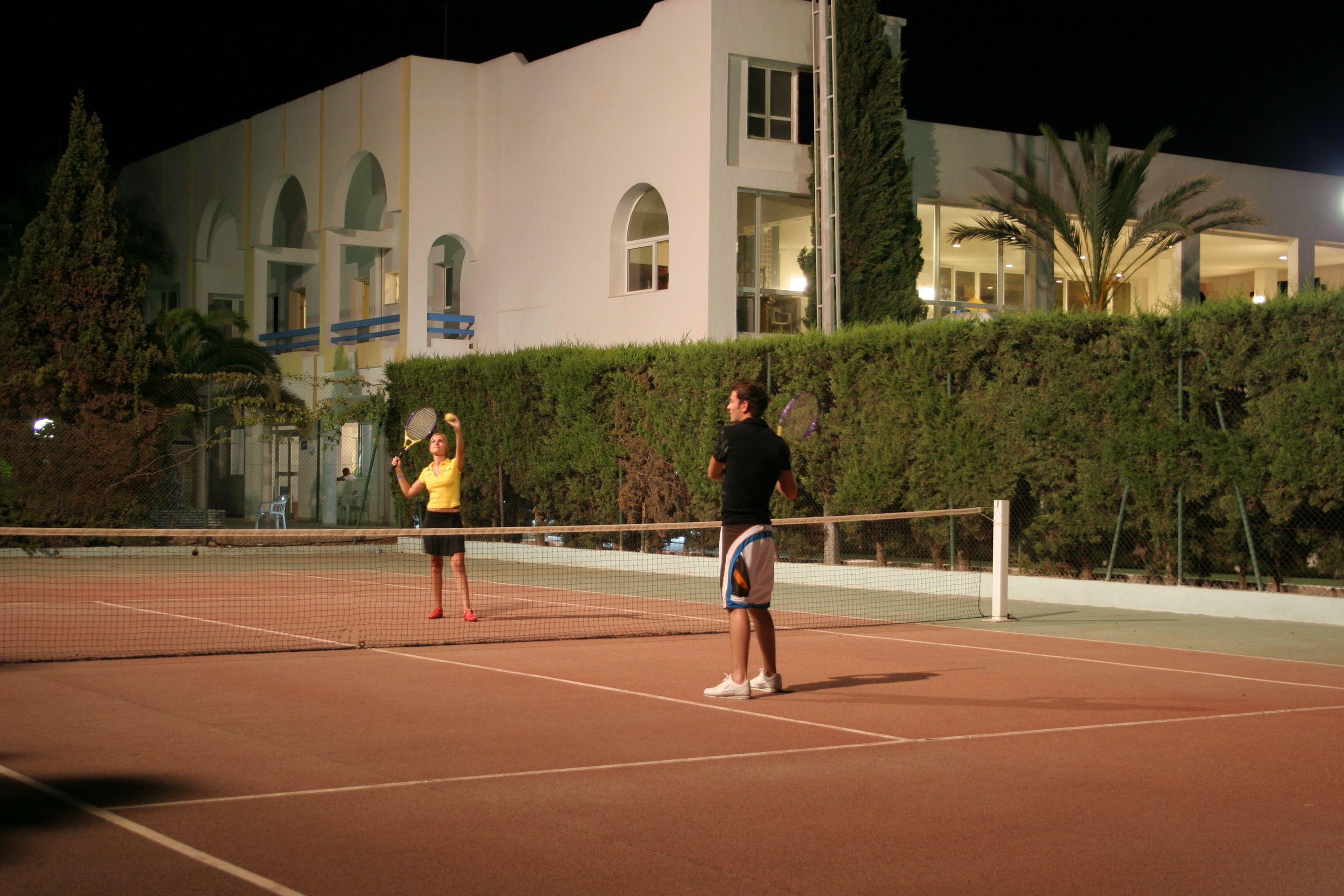 A tennis court with two players, a building with arched windows, and a hedge in the background.