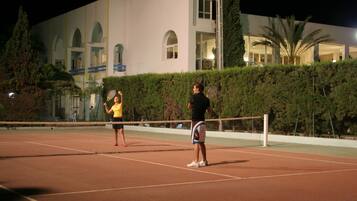 A tennis court with two players, a building with arched windows, and a hedge in the background.
