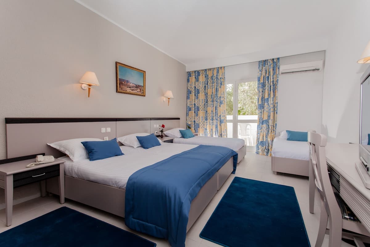 Une chambre d’hôtel avec deux lits, un bureau et une chaise. Il y a un tableau au mur, une fenêtre avec des rideaux et un appareil de climatisation.