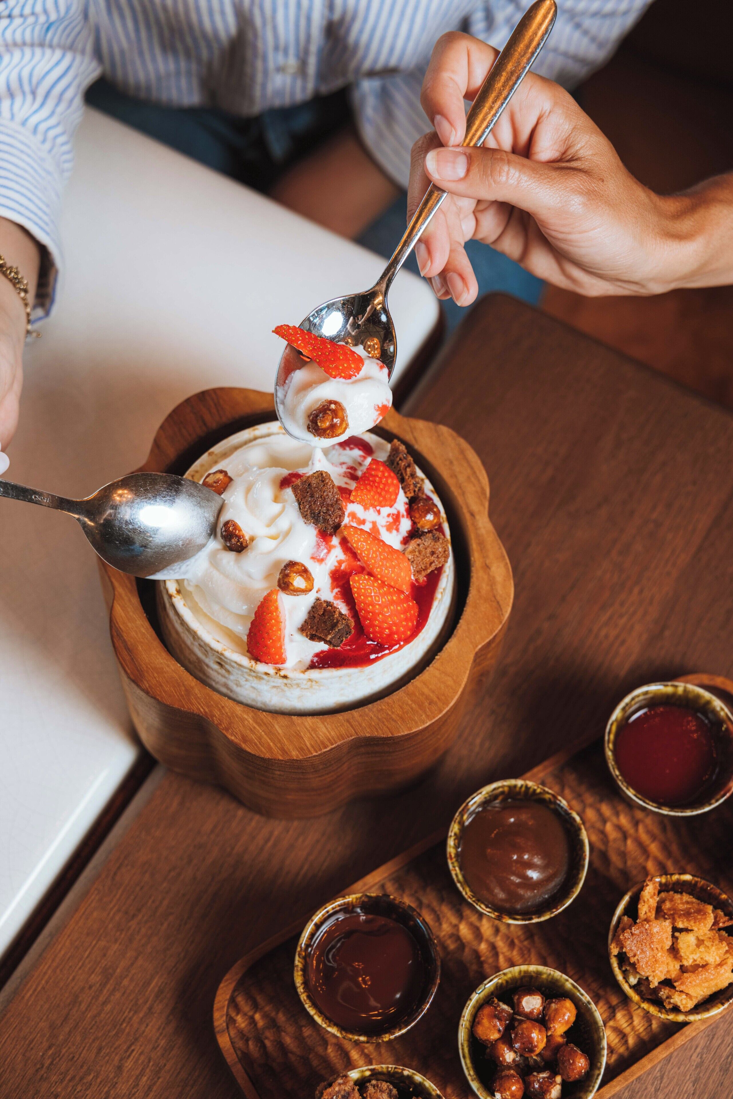 A wooden bowl with a dessert topped with strawberries and nuts, a spoon, and small bowls of sauces on a wooden tray.