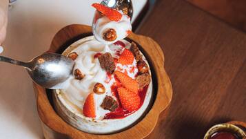 A wooden bowl with a dessert topped with strawberries and nuts, a spoon, and small bowls of sauces on a wooden tray.