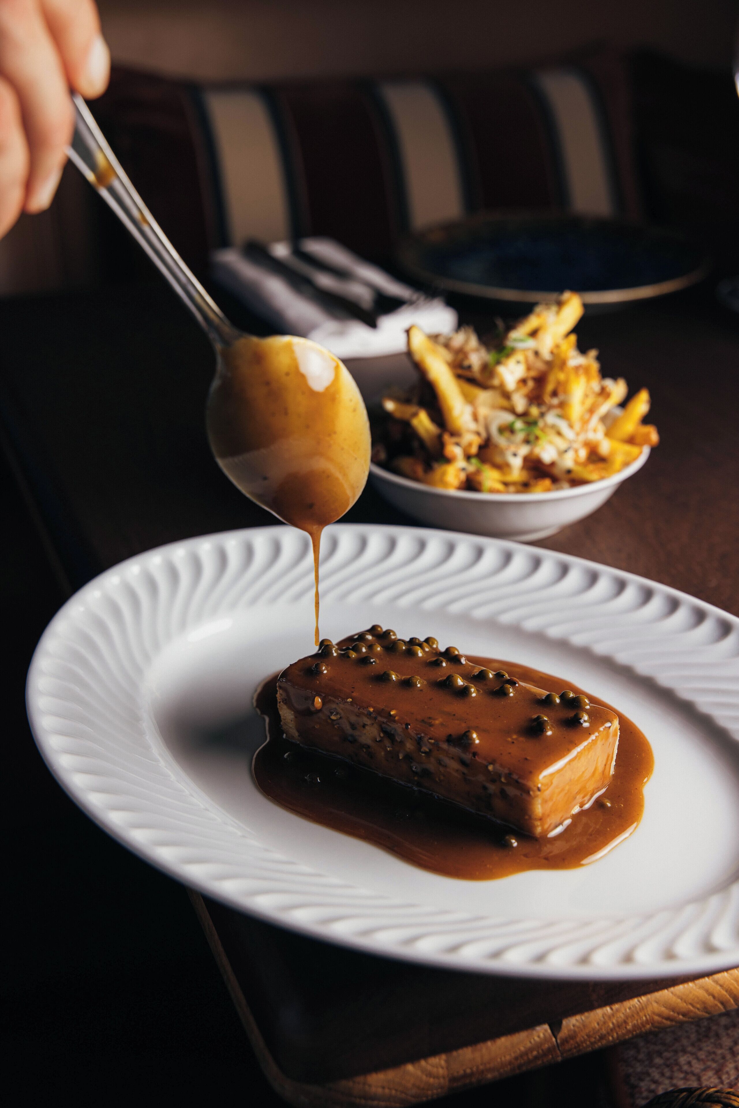 A white plate with a piece of meat topped with sauce and capers, a spoon with sauce, and a bowl of fries in the background.