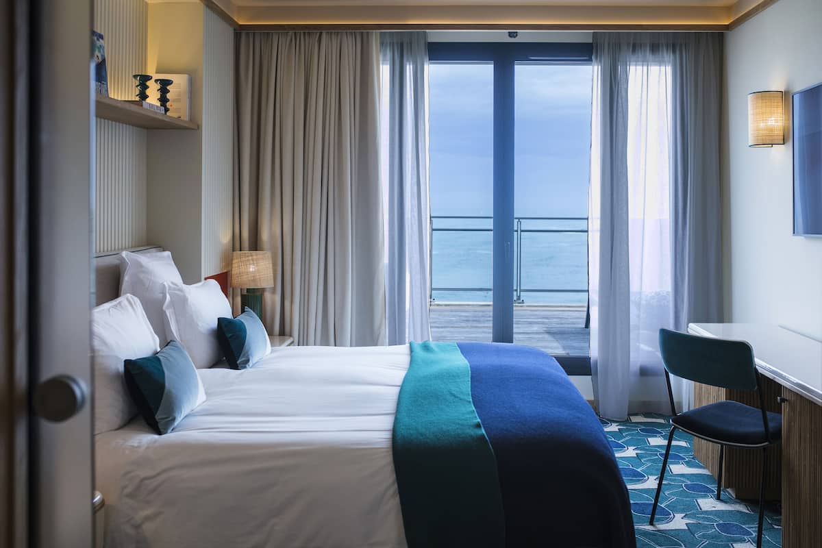 A hotel room with a bed, a desk, a chair, a view of the sea, and a balcony.