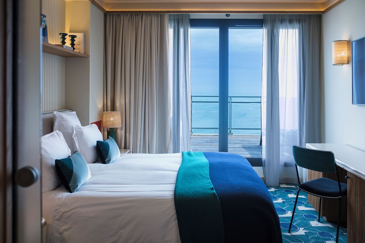 A hotel room with a bed, a desk, a chair, a view of the sea, and a balcony.