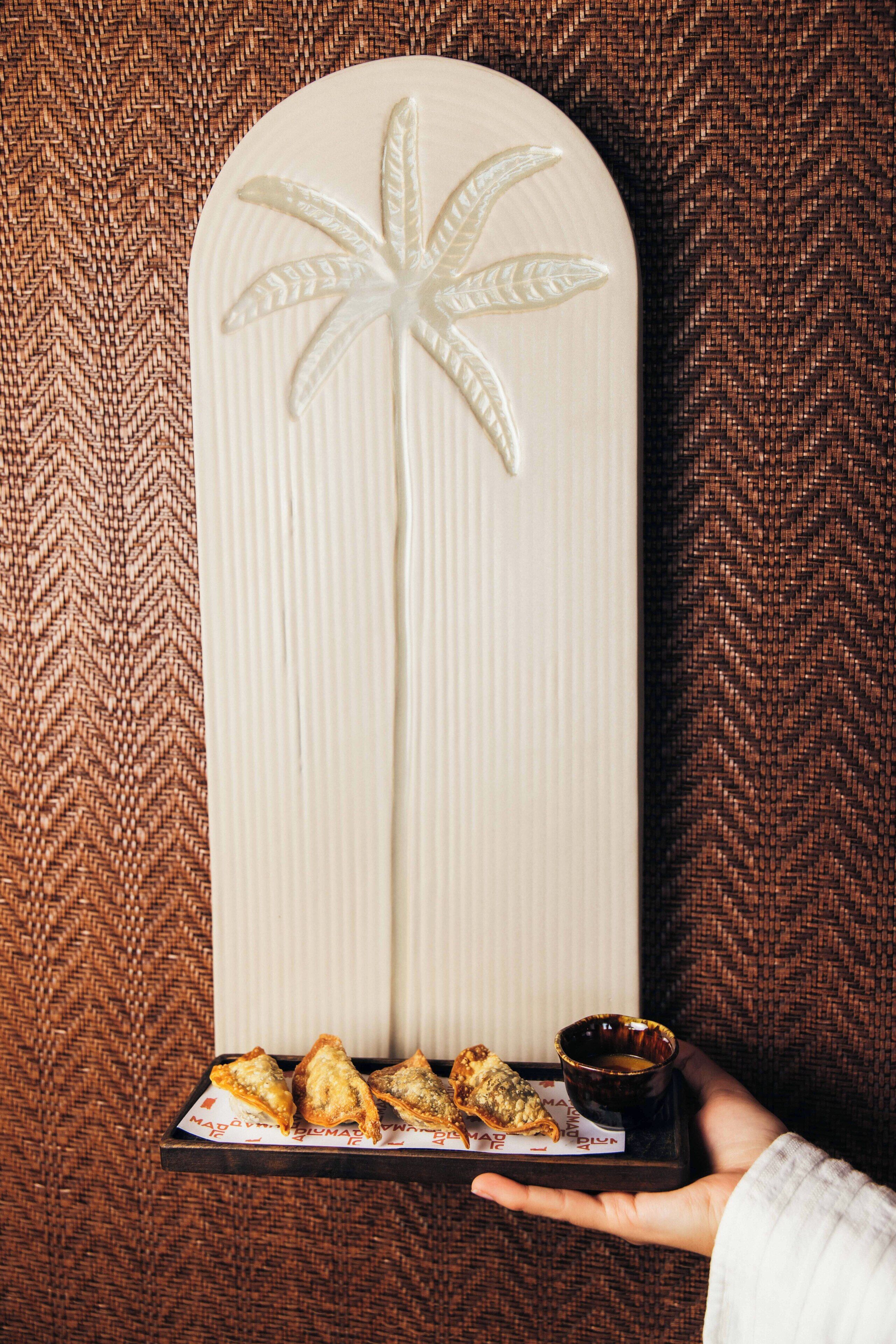 Een hand houdt een dienblad vast met samosa's en een klein kopje, voor een gestructureerde muur.