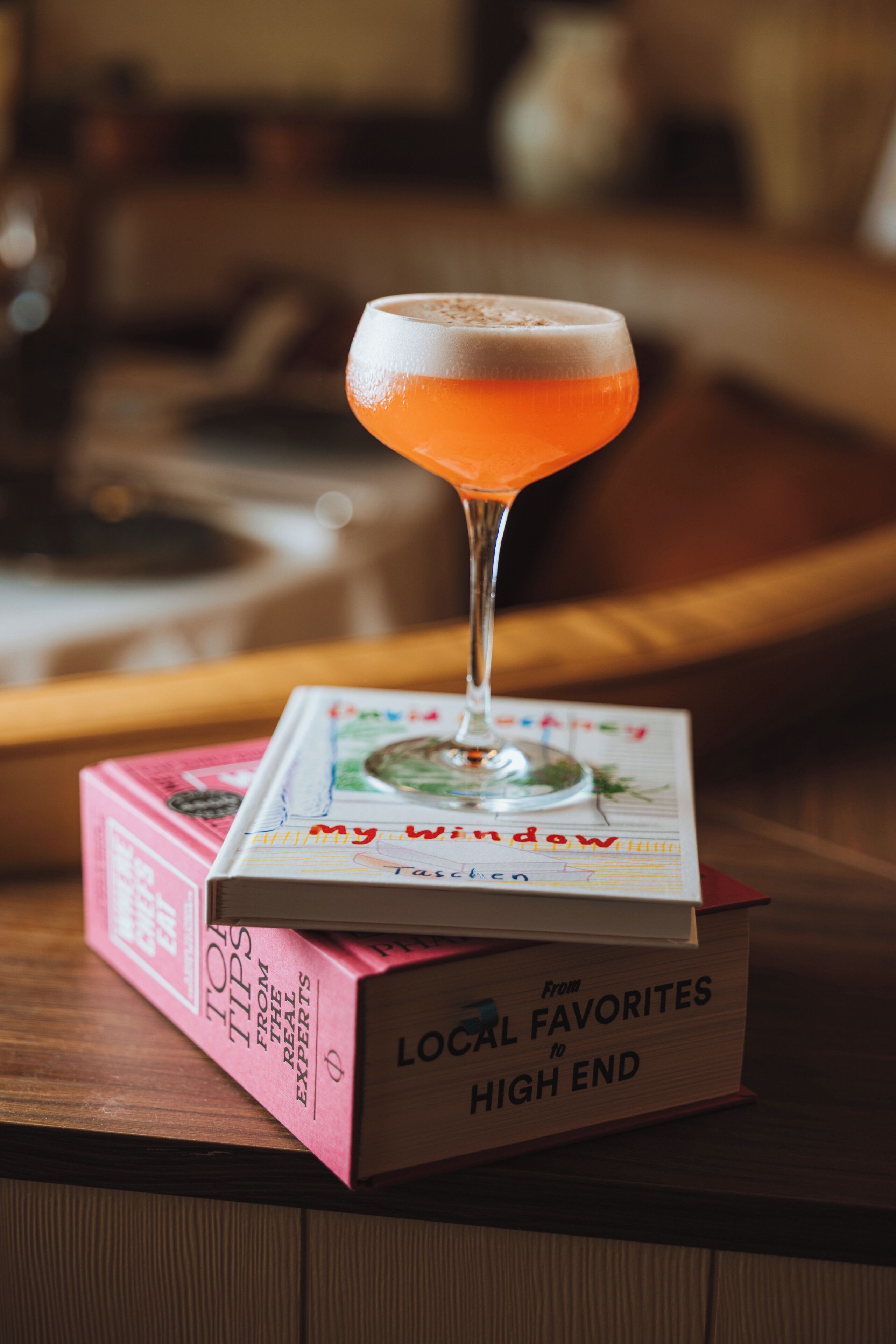 A cocktail glass on a book titled 'My Window Taschen' with a pink box labeled 'Top Tips from the Real Experts' and 'Local Favorites to High End' on a wooden surface.