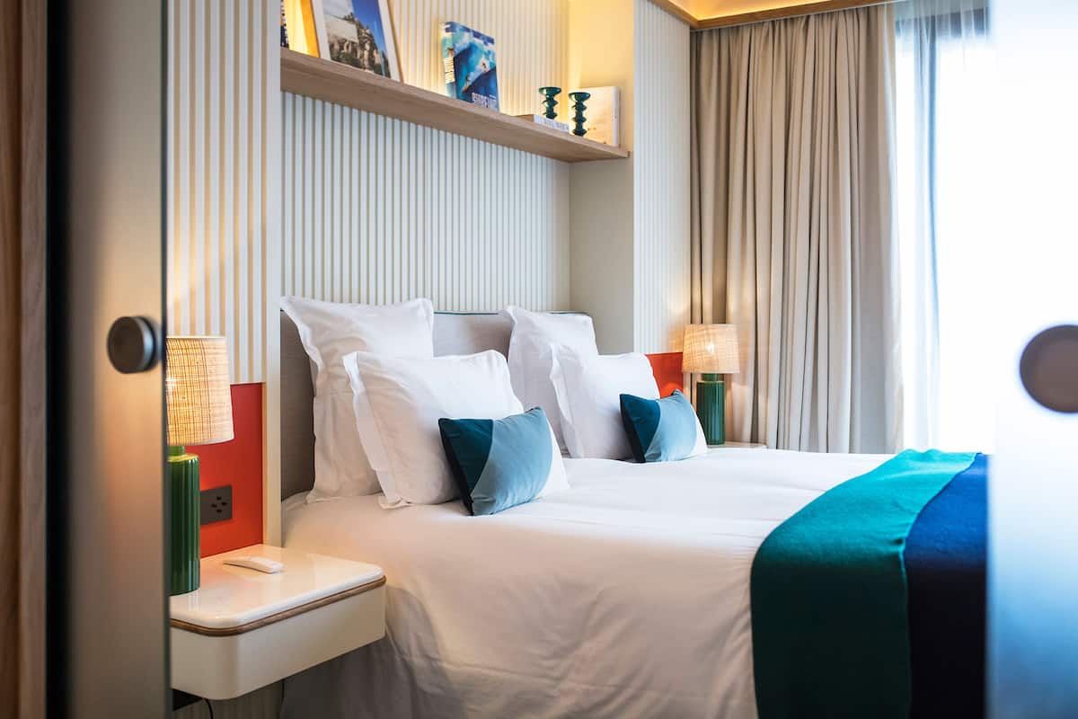 A hotel room with a neatly made bed, bedside lamps, a shelf with books and decorative items, and a window with curtains.