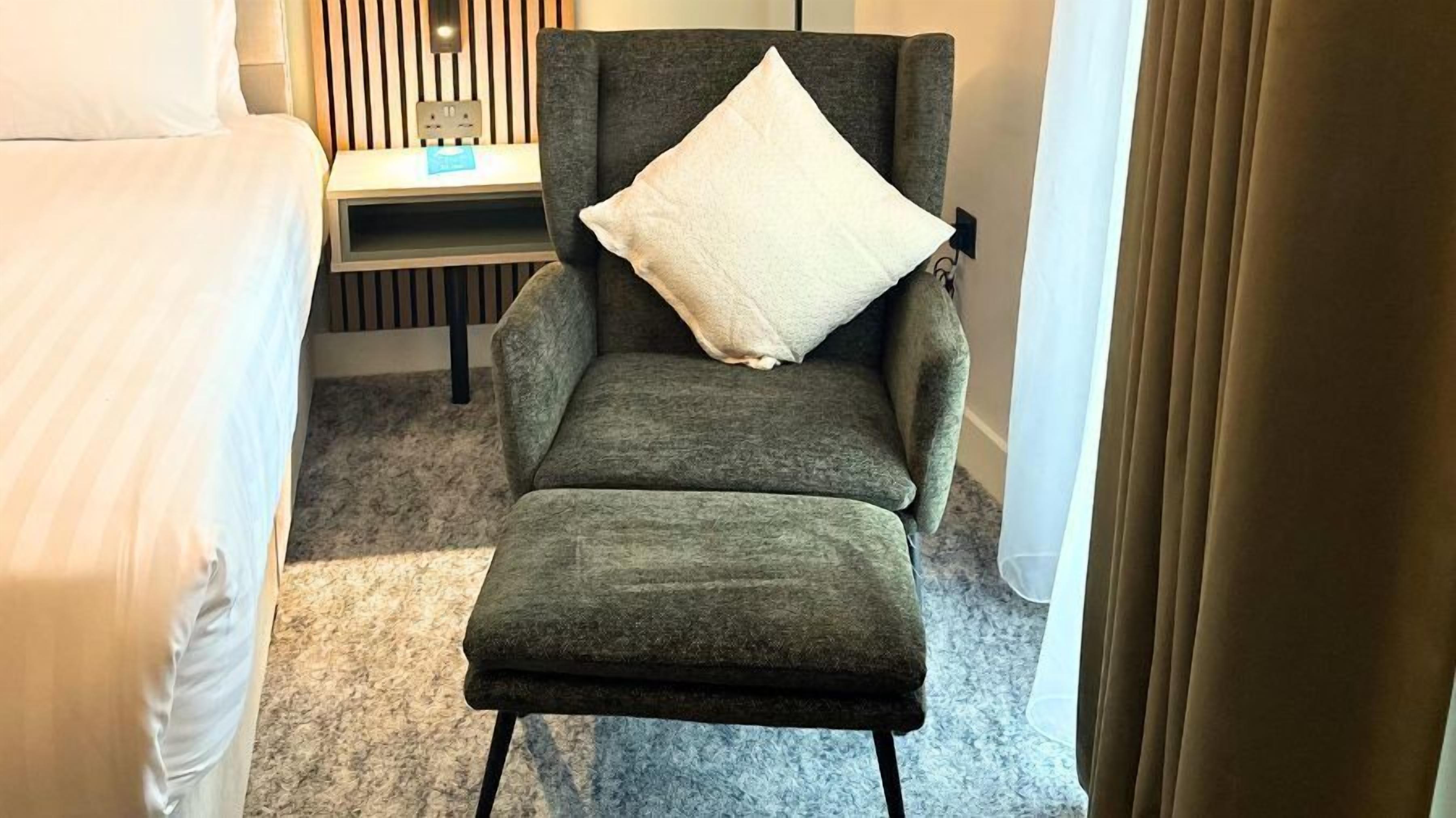 Una habitación de hotel con un sillón gris, una almohada blanca y una mesita con una tarjeta azul.