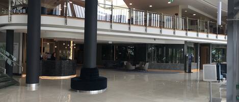 A spacious lobby with a high ceiling, a reception desk, seating area, and a staircase leading to upper floors.