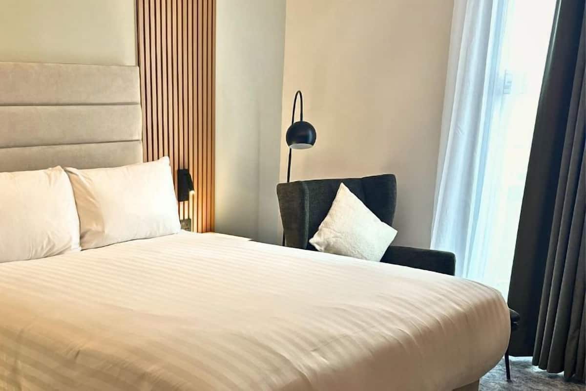 Una cama grande con cabecera, dos almohadas, una mesita de noche con lámpara y una silla con cojín.