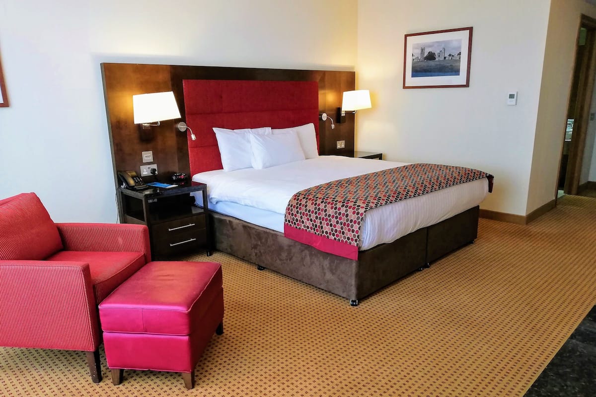 Habitación de hotel con una cama grande, dos mesitas de noche con lámparas, un sillón tapizado rojo y un pequeño otoman rojo.
