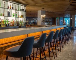 Zona de bar con una barra larga, varios taburetes y una variedad de botellas en estanterías.
