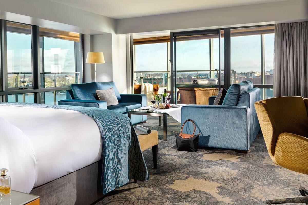 A hotel room with a large bed, a sofa, a dining table, and a view of the city.