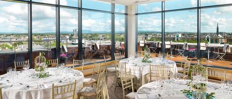 Un área de comedor en la azotea con varias mesas redondas preparadas para un evento formal, ventanas amplias que ofrecen vistas a la ciudad y un cielo despejado.