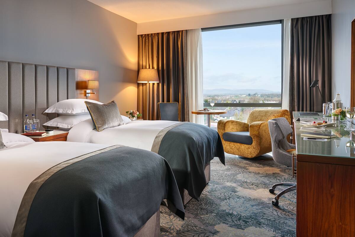 A hotel room with two beds, a desk, a chair, a lamp, and a view of the city.