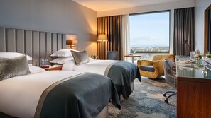 A hotel room with two beds, a desk, a chair, a lamp, and a view of the city.