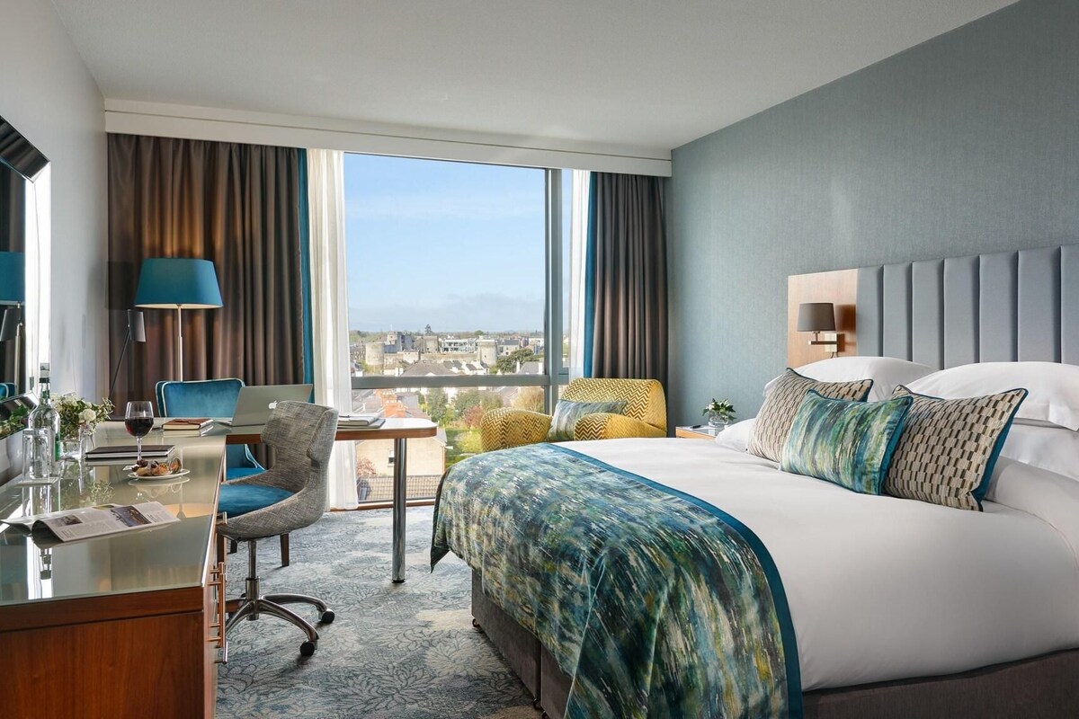 A hotel room with a large bed, a desk with a laptop, a chair, and a view of the city.