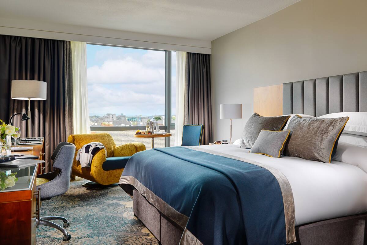 A hotel room with a large bed, a desk with a chair, a window with curtains, and a view of the city.