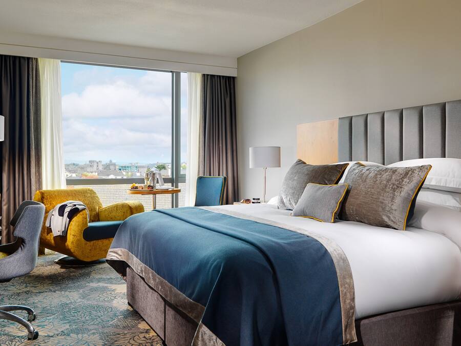 Una habitación de hotel con una cama grande, un escritorio con silla, una ventana con cortinas y vista a la ciudad.
