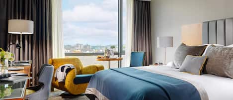 A hotel room with a large bed, a desk with a lamp, a chair, and a view of the city through a large window.