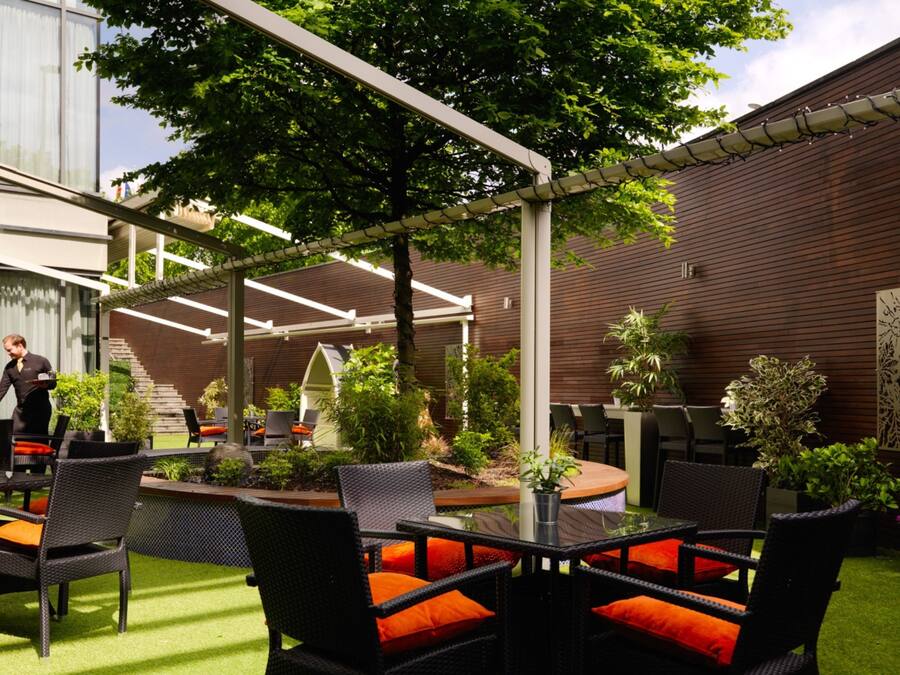 An outdoor patio area with wicker furniture and orange cushions, surrounded by greenery and a wooden wall.