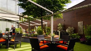 Un patio al aire libre con mobiliario de mimbre y cojines naranja, rodeado de follaje y un muro de madera.