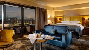 Camera d'albergo con un letto grande, un divano, un tavolo da pranzo con una candela e vista sulla città attraverso ampie finestre.