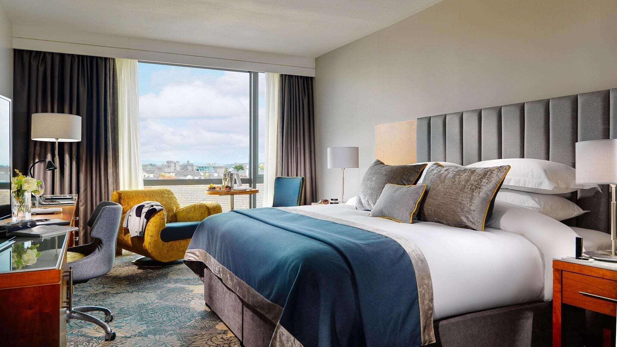 A hotel room with a large bed, a desk, a chair, and a view of the city.