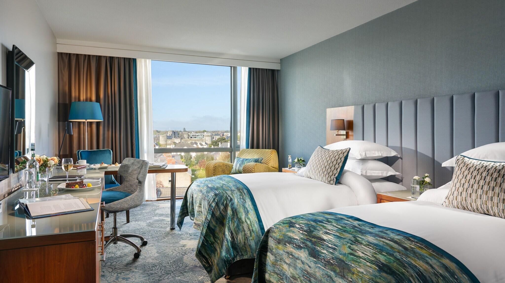A hotel room with a large bed, a desk with a chair, a lamp, and a view of the city.