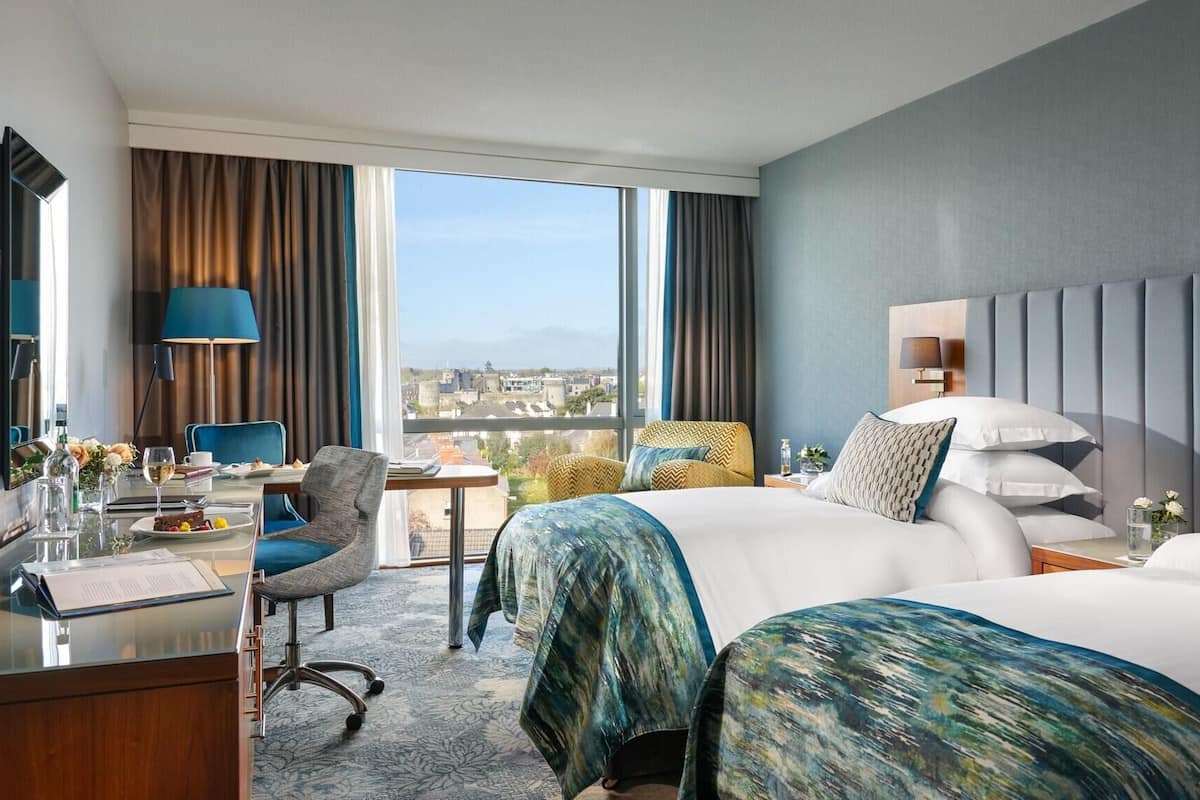 A hotel room with a large bed, a desk with a chair, a lamp, and a view of the city.