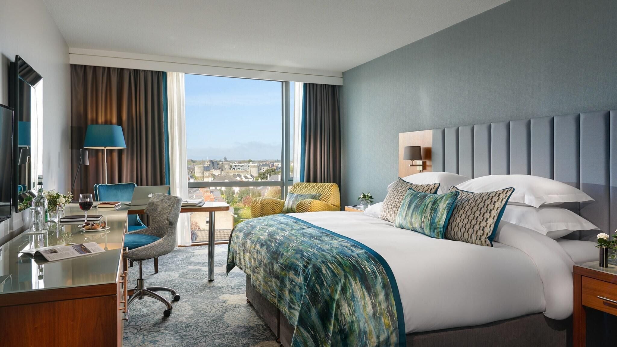 A hotel room with a large bed, a desk with a laptop, a chair, and a view of the city.