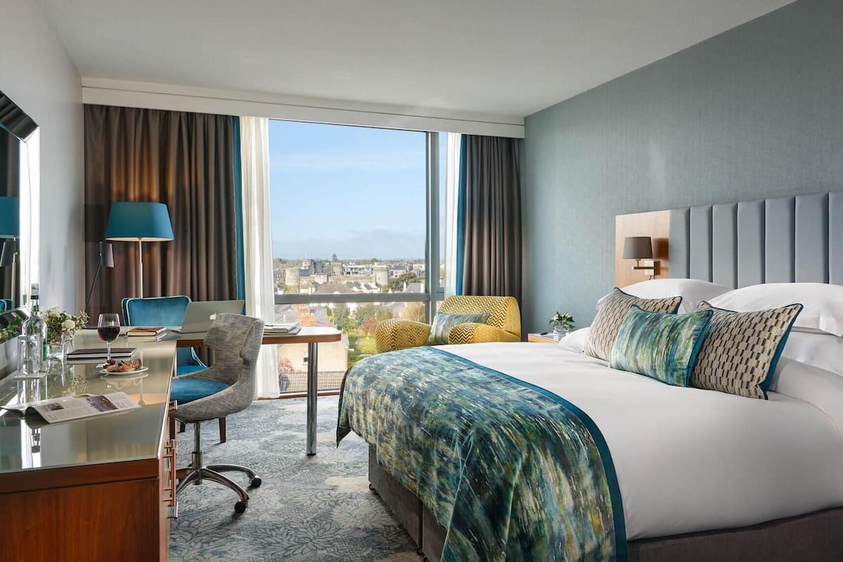 A hotel room with a large bed, a desk with a laptop, a chair, and a view of the city.