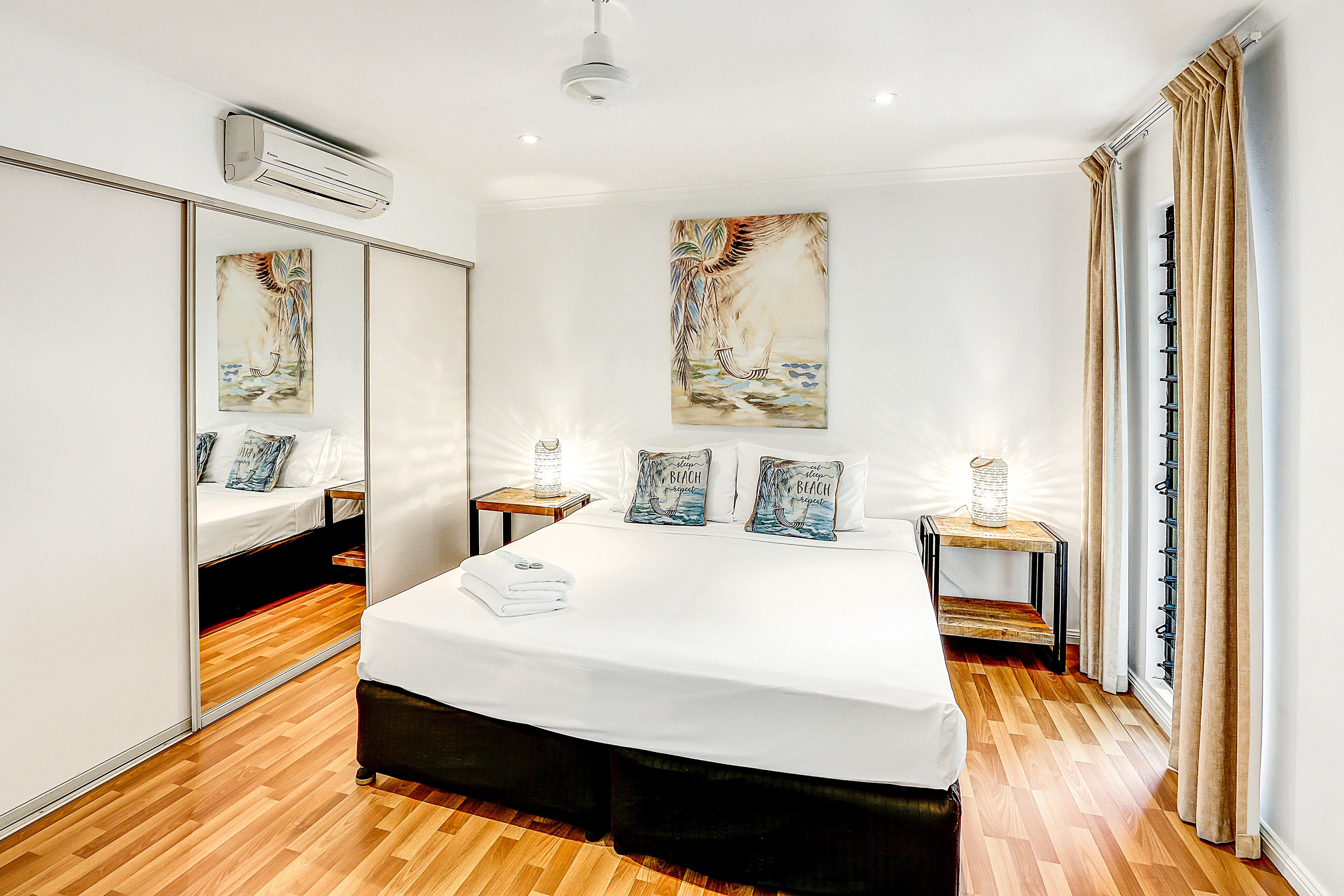 Quarto de hotel com uma cama grande, mesa de cabeceira com abajur, espelho e obra de arte na parede.