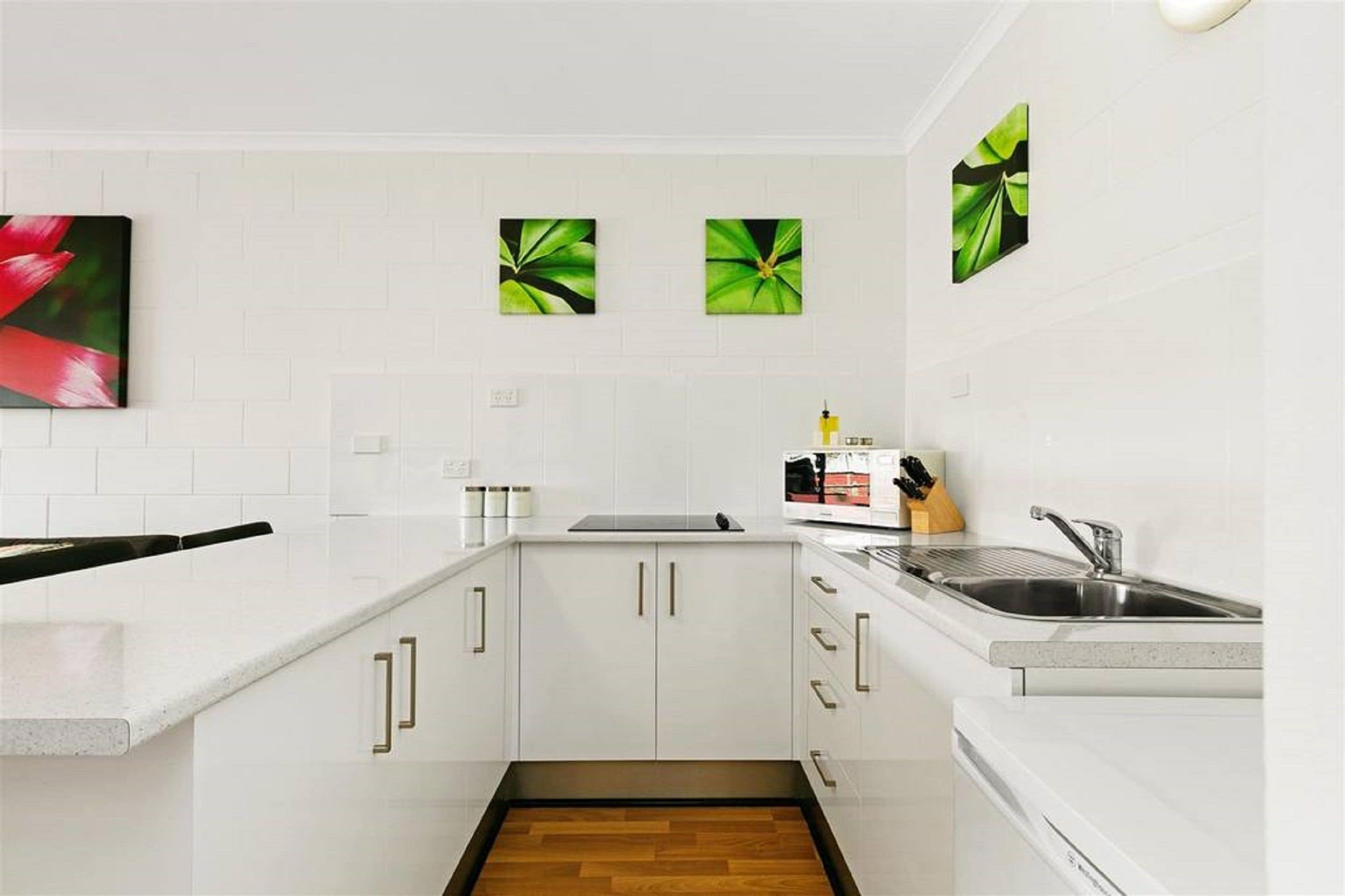 A modern kitchen with white cabinets, a stainless steel sink, and a microwave. There are three green leaf paintings on the wall.