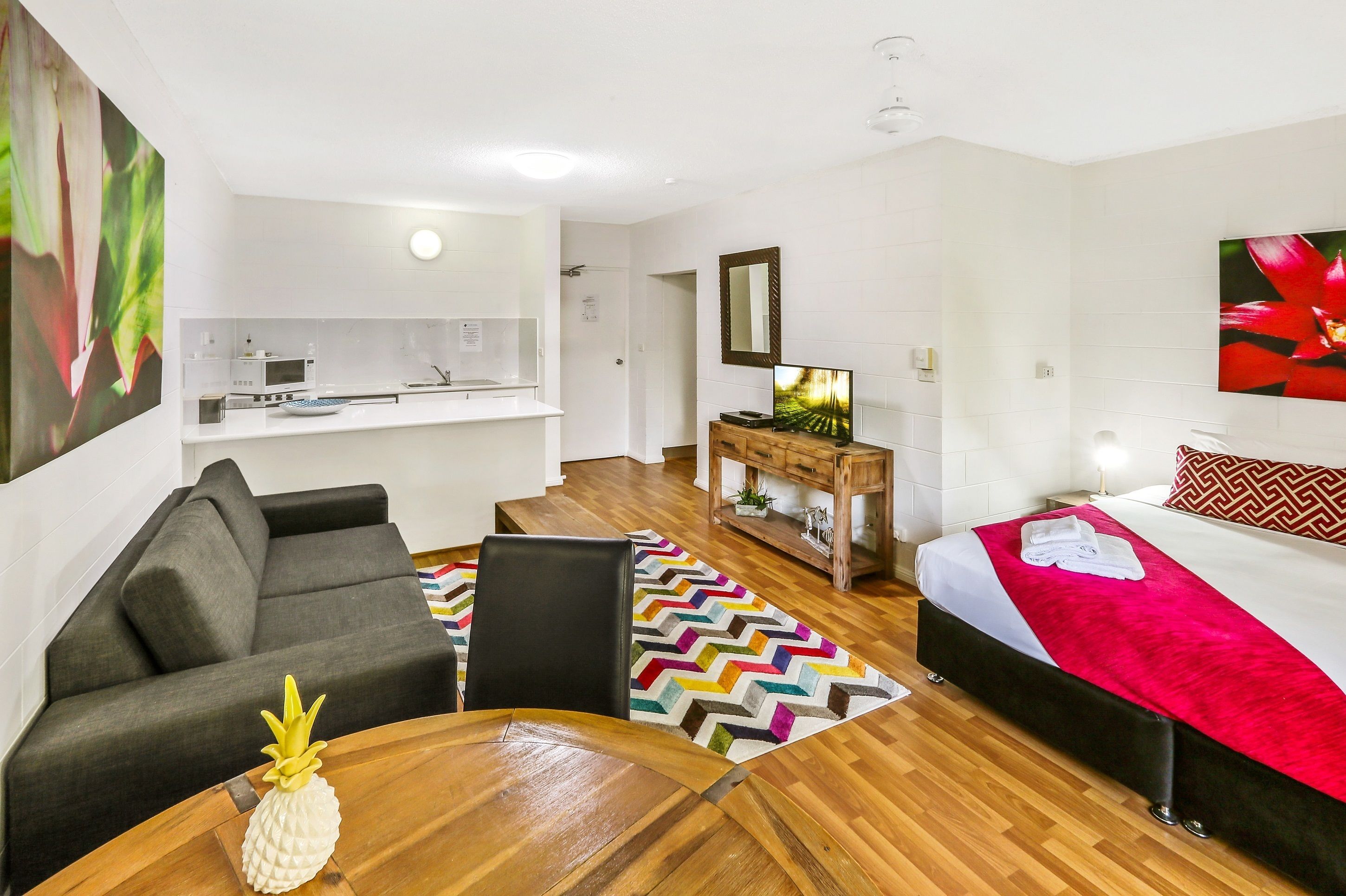 Une chambre d’hôtel moderne avec un canapé, une table à manger, un lit recouvert d’une couverture rouge, une commode en bois et, au fond, un coin cuisine.
