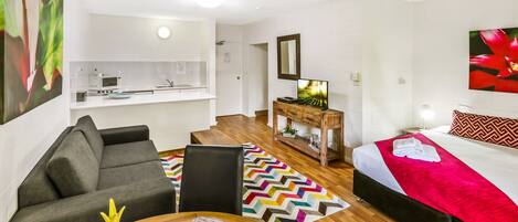 Une chambre d’hôtel moderne avec un canapé, une table à manger, un lit recouvert d’une couverture rouge, une commode en bois et, au fond, un coin cuisine.