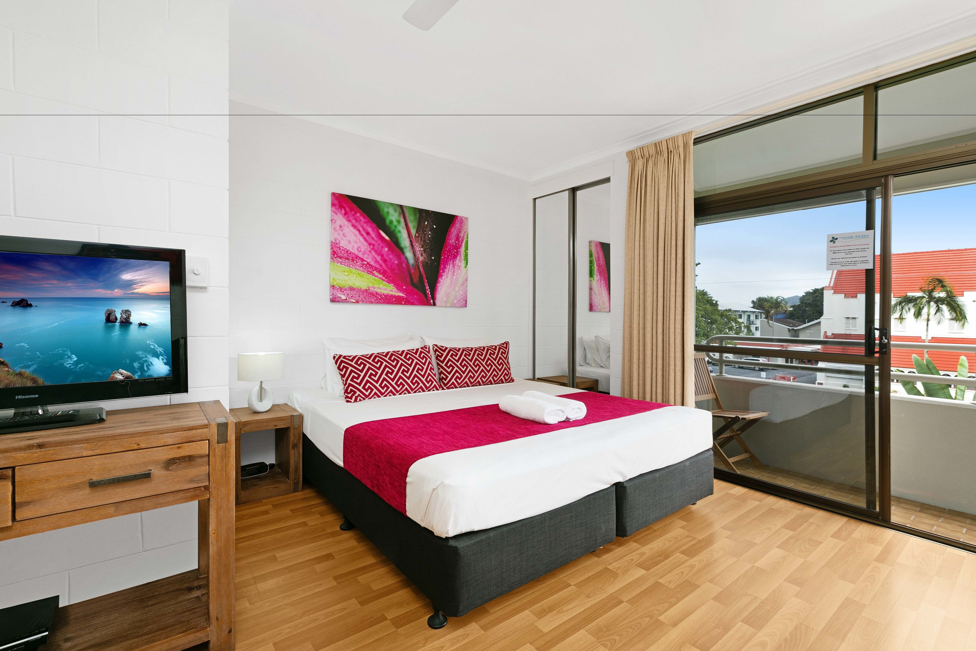 A modern hotel room with a large bed, a flat-screen TV, a wooden dresser, and a balcony with a view of the city.