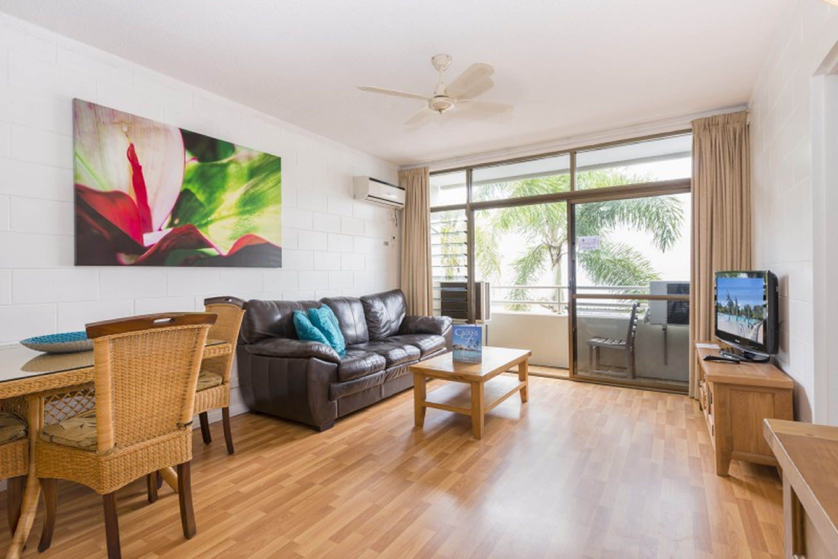 A modern living room with a black leather sofa, wicker chairs, a wooden coffee table, a flat-screen TV, and a large window with a view of palm trees.