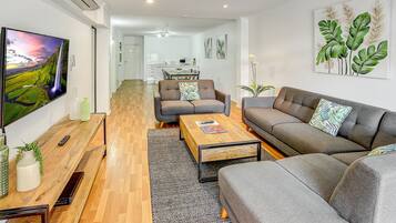 A modern living room with a flat-screen TV, grey sofas, a wooden coffee table, and a kitchen in the background.