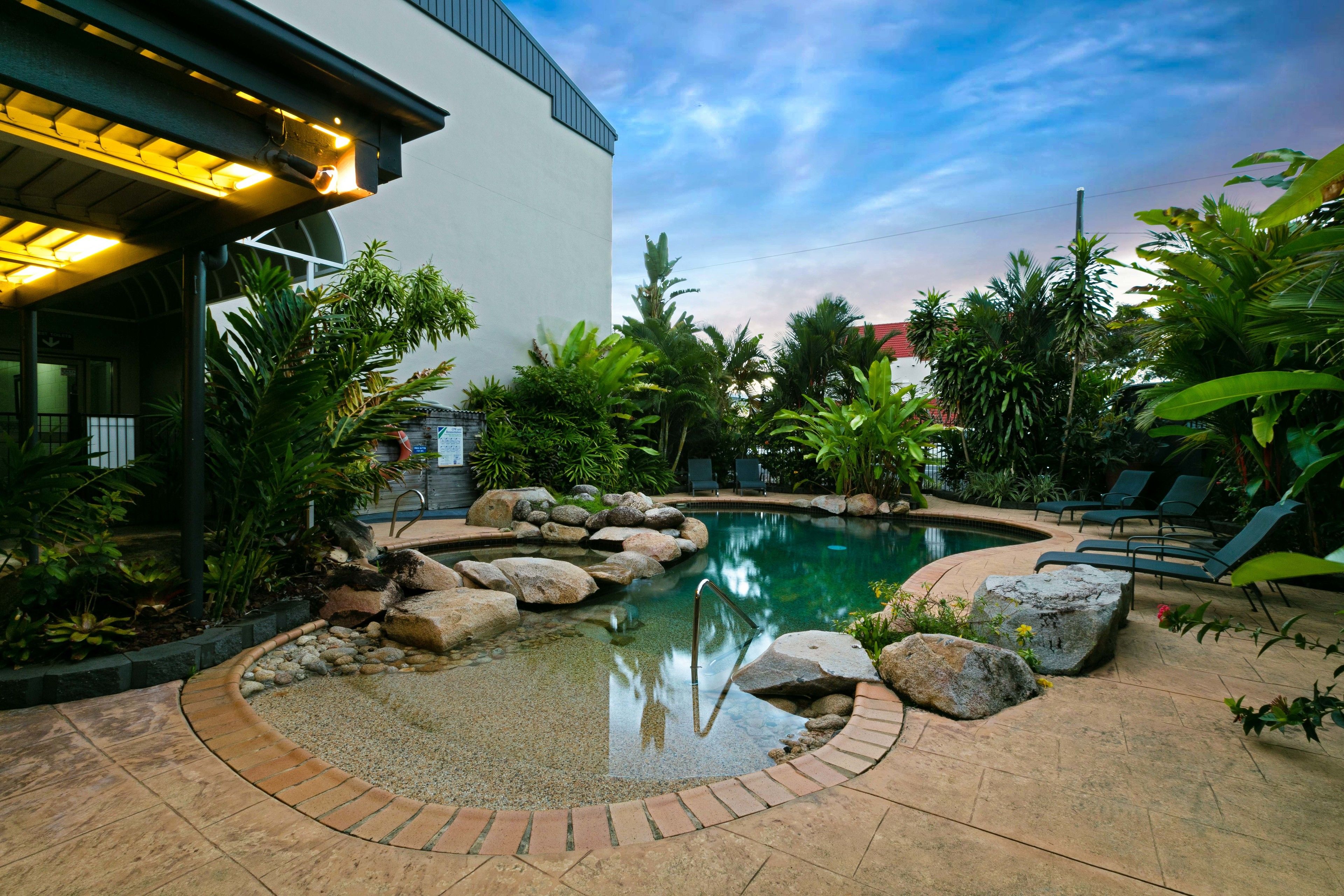 A swimming pool with rocks and plants, surrounded by a brick patio and lounge chairs.
