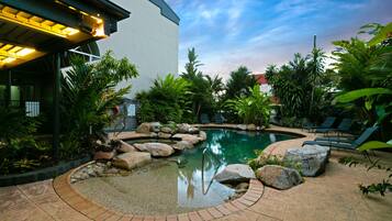 A swimming pool with rocks and plants, surrounded by a brick patio and lounge chairs.