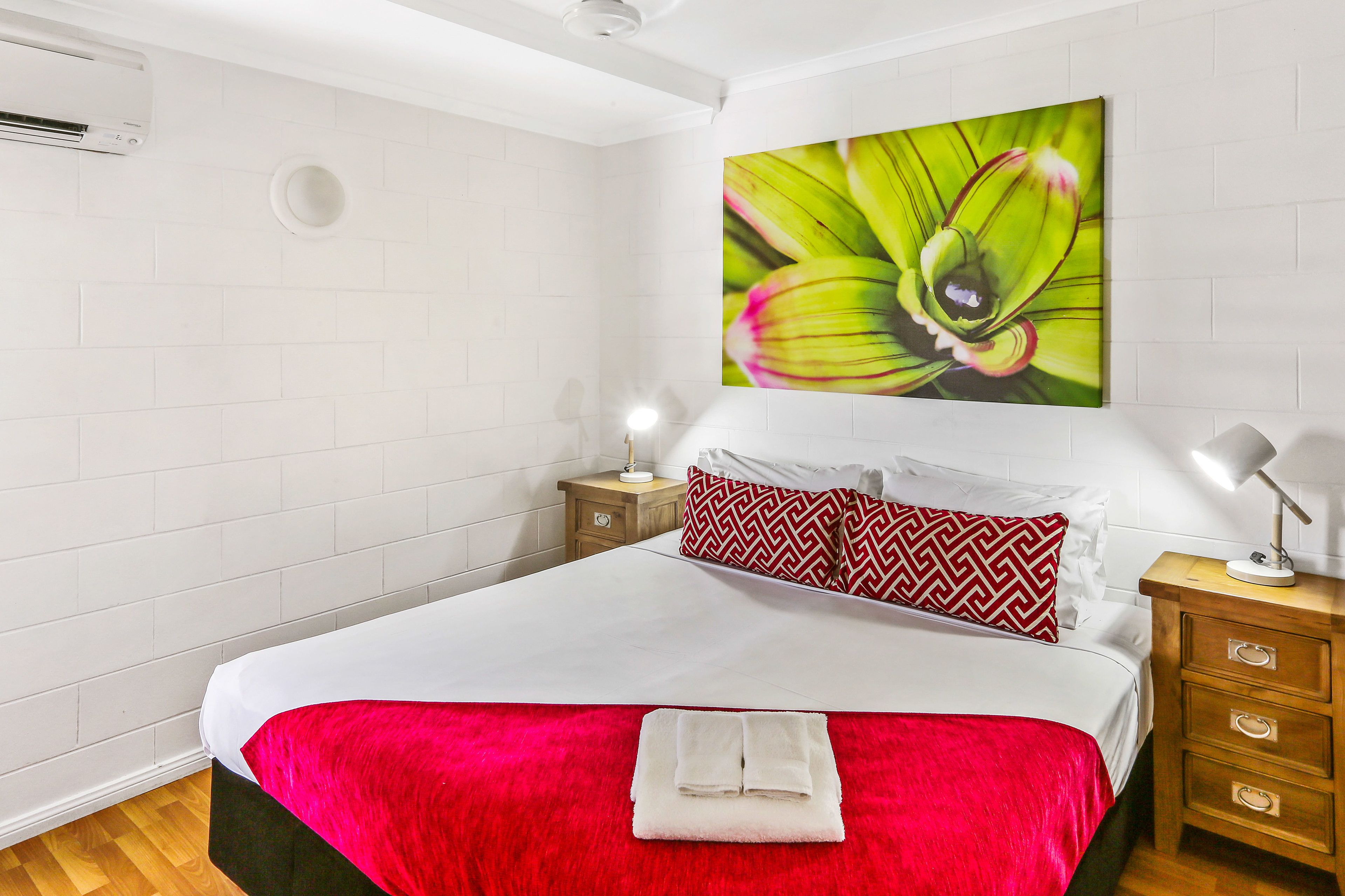 Habitación de hotel con una cama grande, paredes blancas, un cojín con estampado rojo y blanco, un equipo de aire acondicionado montado en la pared y una mesita de noche con una lámpara.
