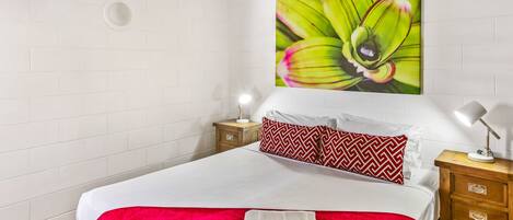Habitación de hotel con una cama grande, paredes blancas, un cojín con estampado rojo y blanco, un equipo de aire acondicionado montado en la pared y una mesita de noche con una lámpara.