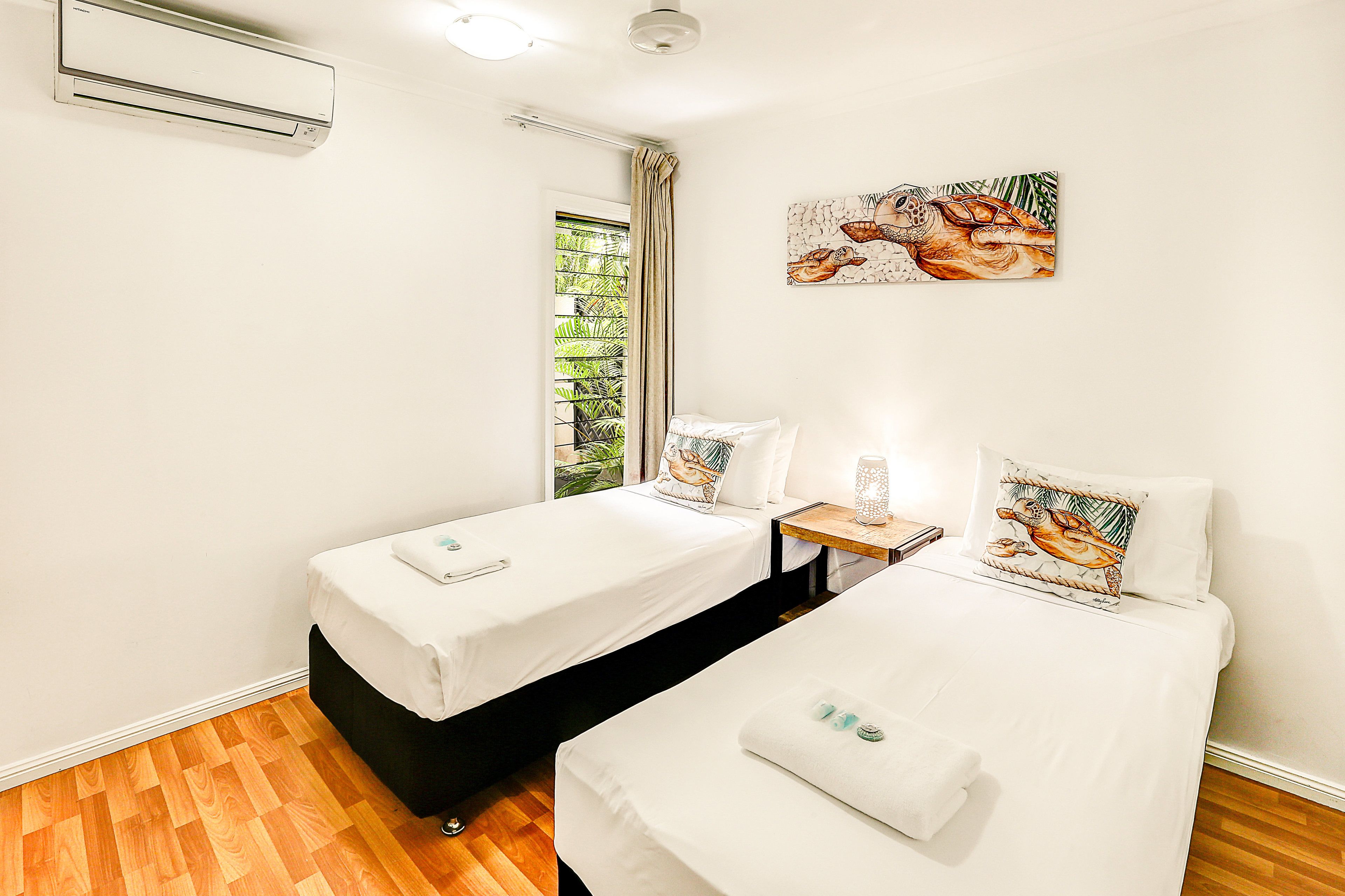 A hotel room with two beds, a window with curtains, a small table with a lamp, and a wall-mounted air conditioner.