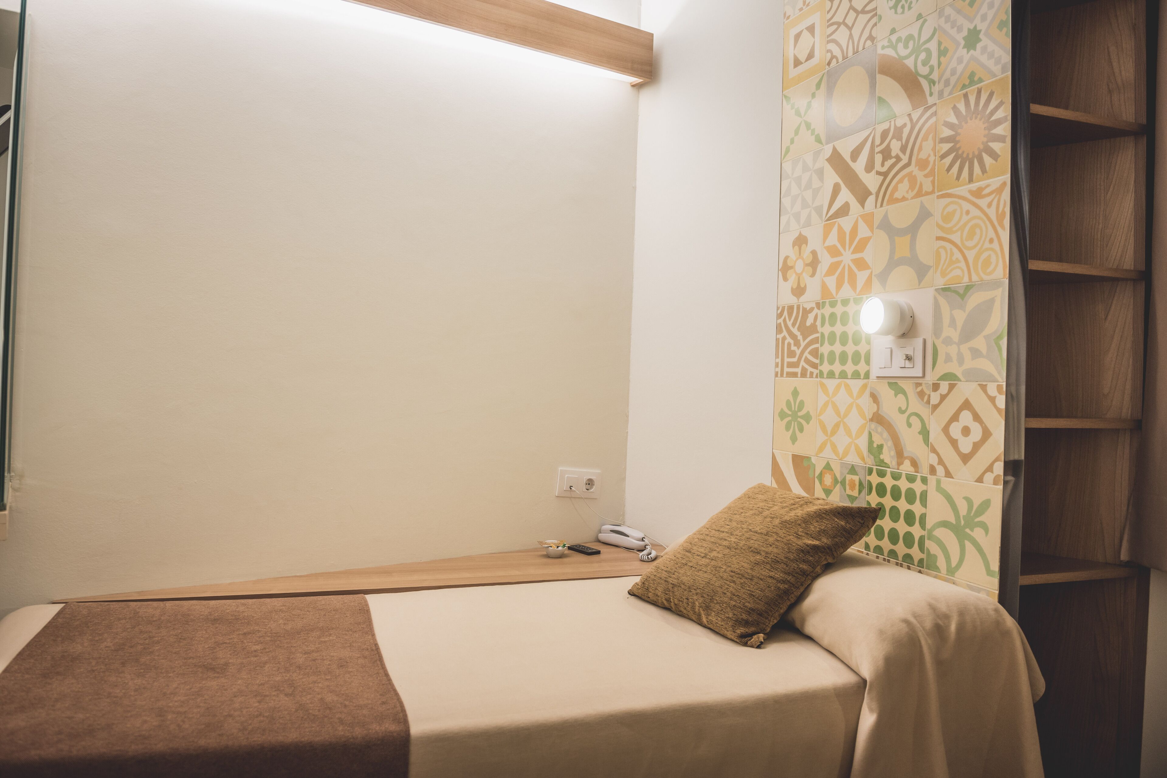 Un petit lit bien fait, avec une couverture et un oreiller marron, une table de chevet sur laquelle se trouvent un téléphone et une télécommande, et un mur recouvert de carreaux décoratifs.
